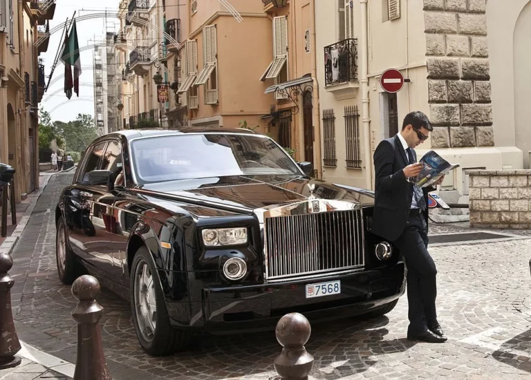 Chauffeur stands by black Rolls Royce car reading magazine