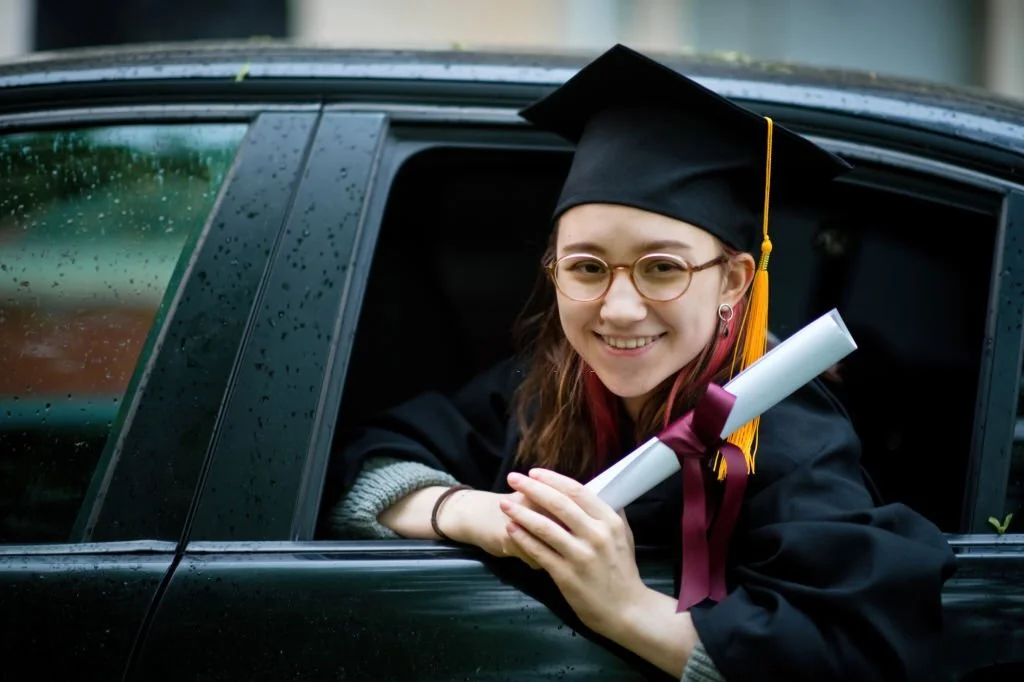 Teenage girl wearing graduation gown and cap with diploma in car